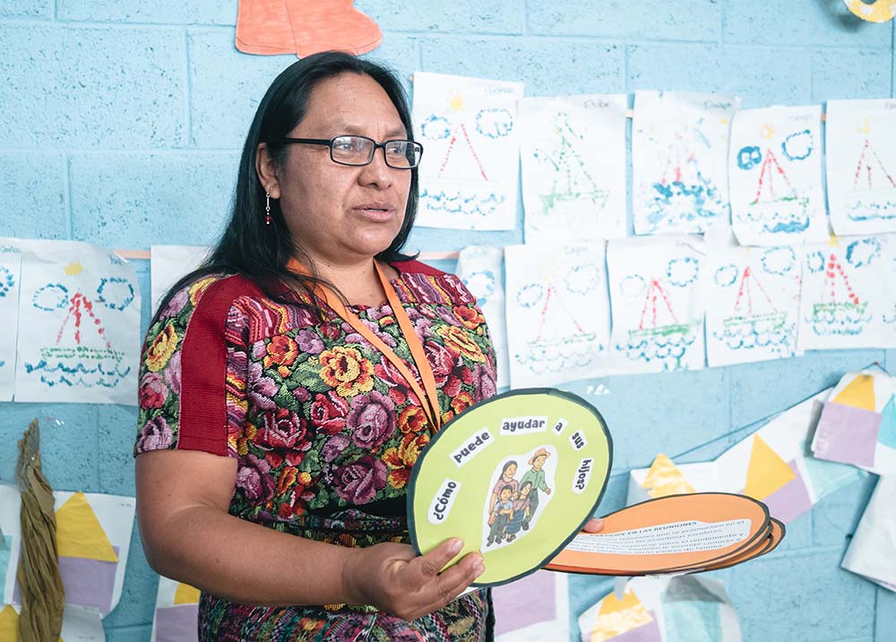 A woman in a floral dress is speaking inside a classroom. She's holding a sign that says "¿Cómo puede ayudar a sus hijos?"